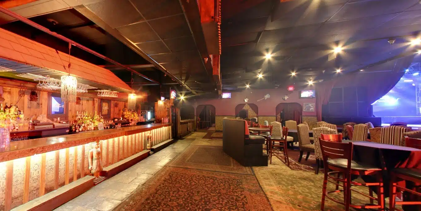 A dimly lit bar in Westlake Village with a long counter on the left, tables and chairs on the right, ornate rugs on the floor, and warm, ambient lighting throughout the room—offering an inviting atmosphere reminiscent of upscale lounges in Agoura Hills.
