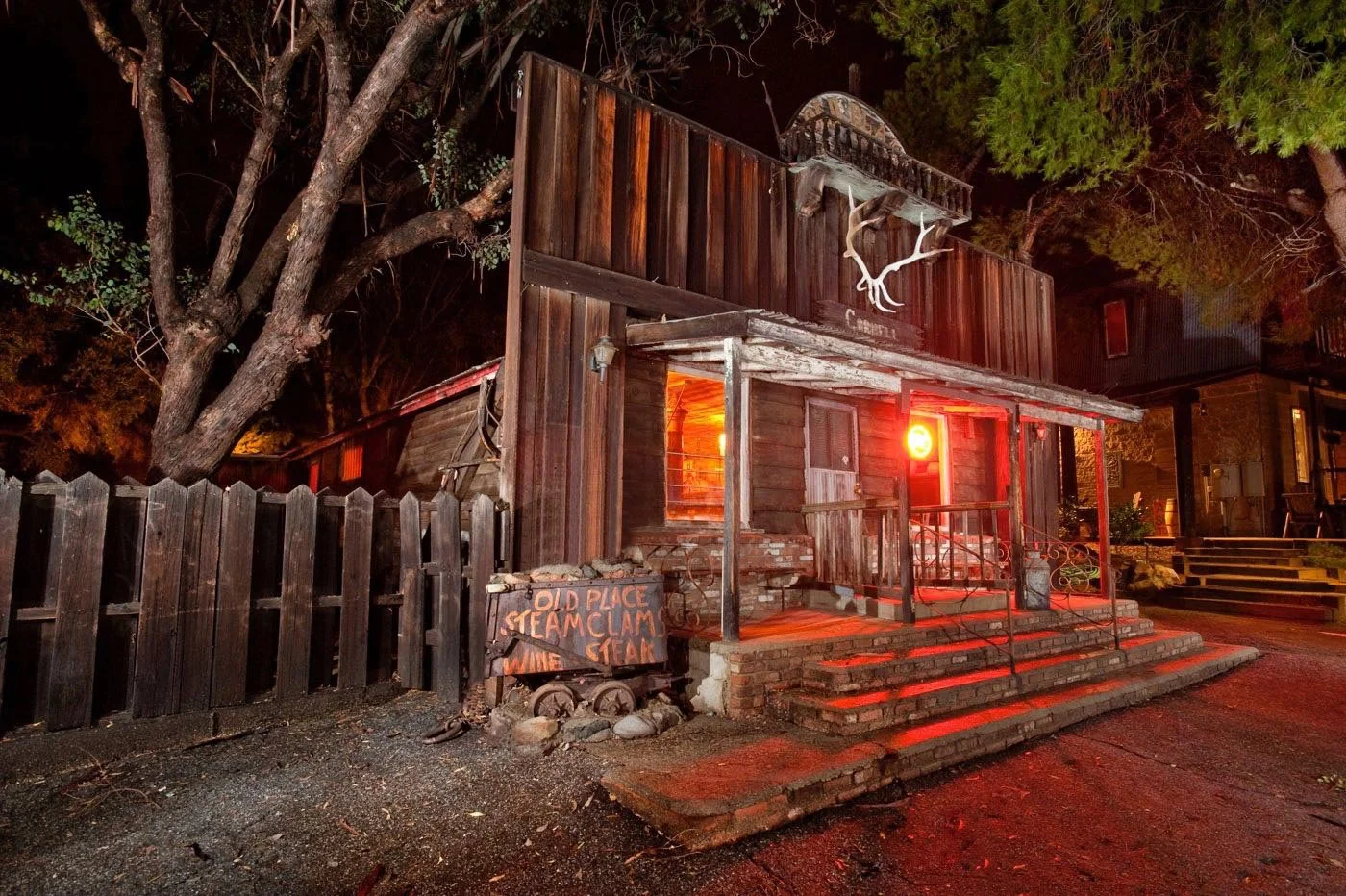 A rustic wooden building at night in Agoura Hills features a glowing red neon light, antlers above the door, and a sign reading “Old Place: Steak, Clam, Wine.” There are trees, a fence, and wooden steps leading to the porch.