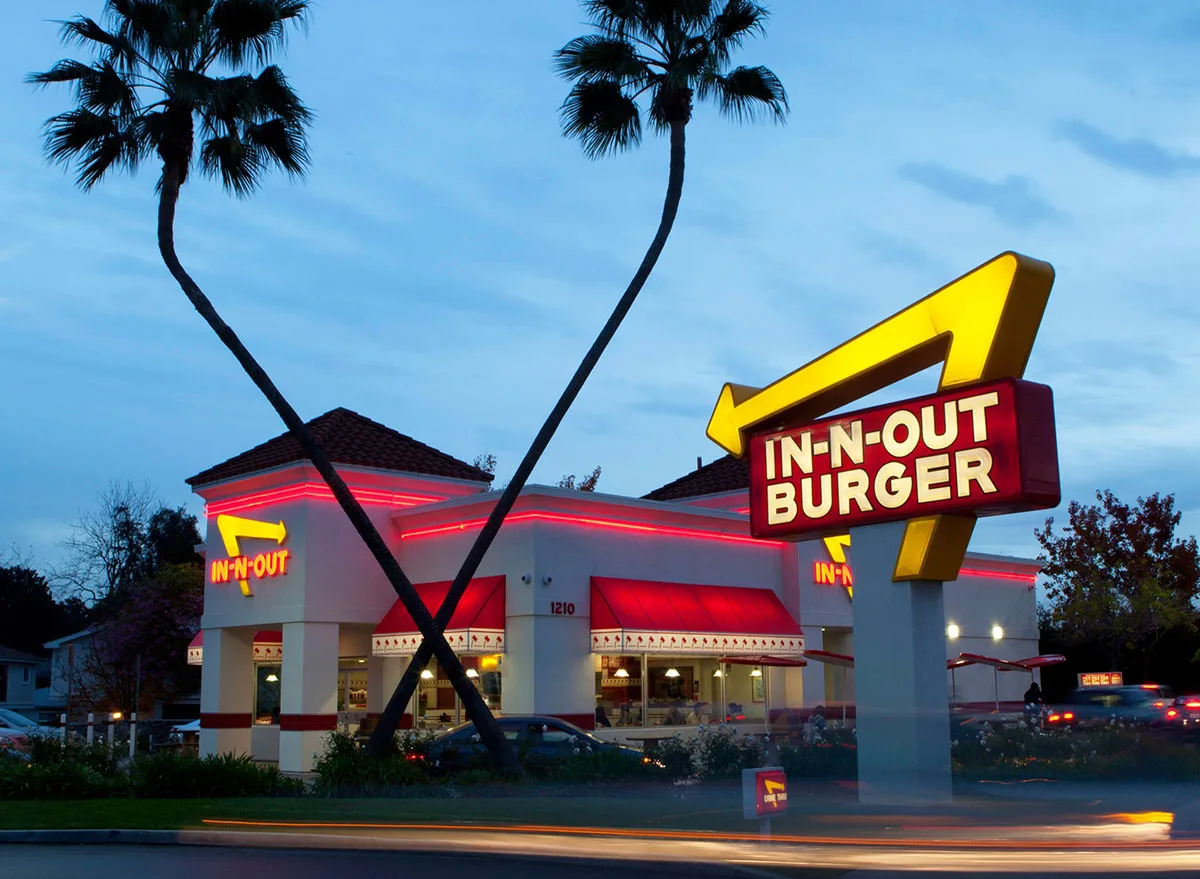 In-N-Out Burger restaurant at dusk in Westlake Village, with illuminated yellow and red signage, palm trees, and cars passing by, creating light trails in the foreground.