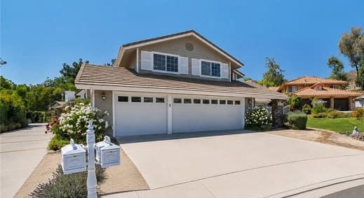 Two-story suburban house with beige siding, brown roof, and three-car garage located at 869 Admiral Court in Oak Park. Two white mailboxes are at the end of the wide driveway. The yard is landscaped with bushes, flowers, and a few trees.